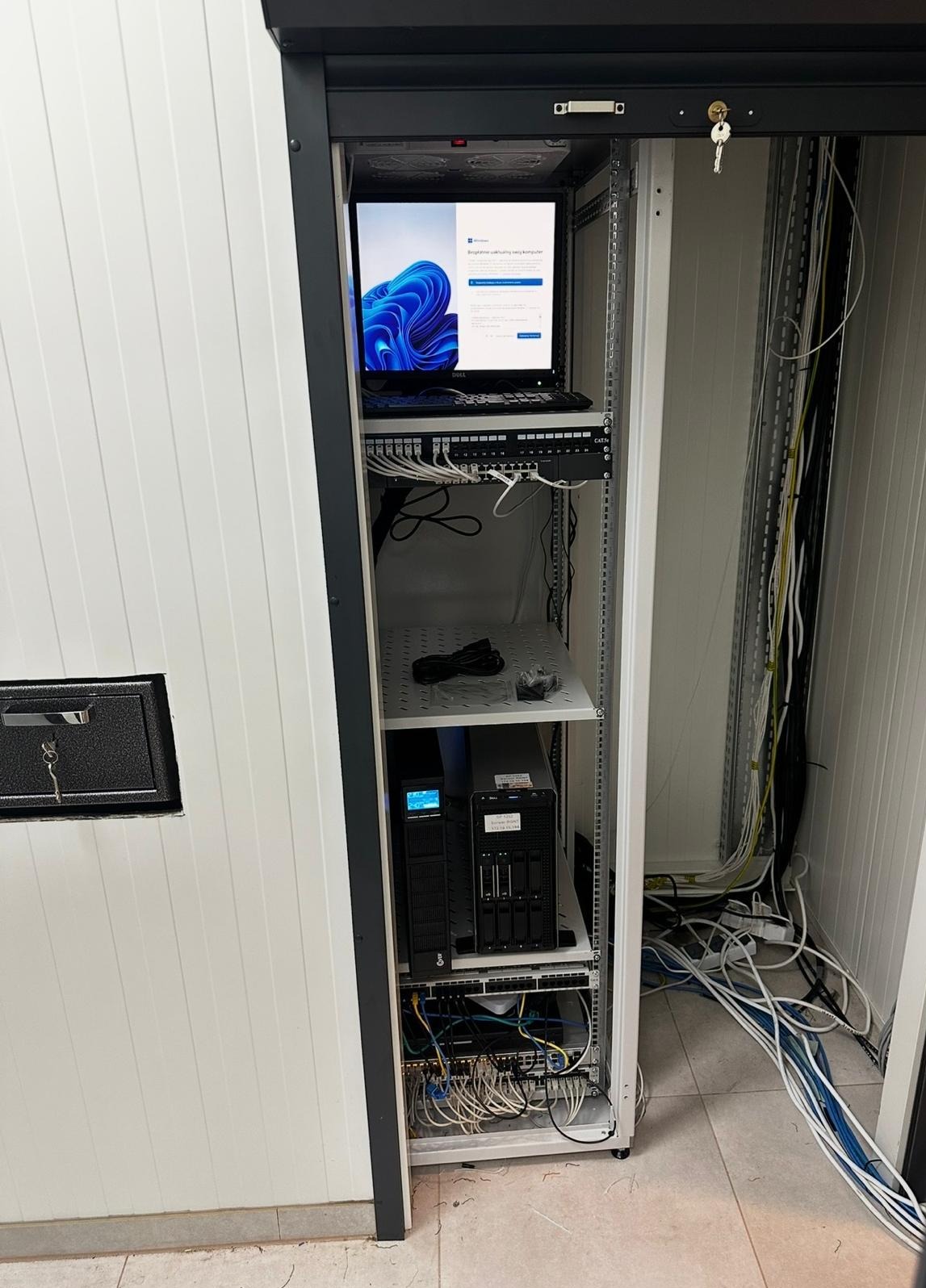 Szafa rack z serwerem, UPS i switchem, laptop z ekranem logowania Windows 11 na górze. Widoczne okablowanie i zabezpieczenia szafy. Instalacja sieci komputerowej w serwerowni.