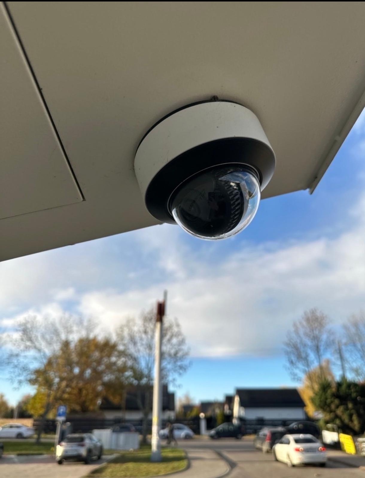 Kamera kopułkowa monitoringu, zamontowana na zewnątrz budynku, z widocznym otoczeniem: drzewa, niebo i fragment drogi.