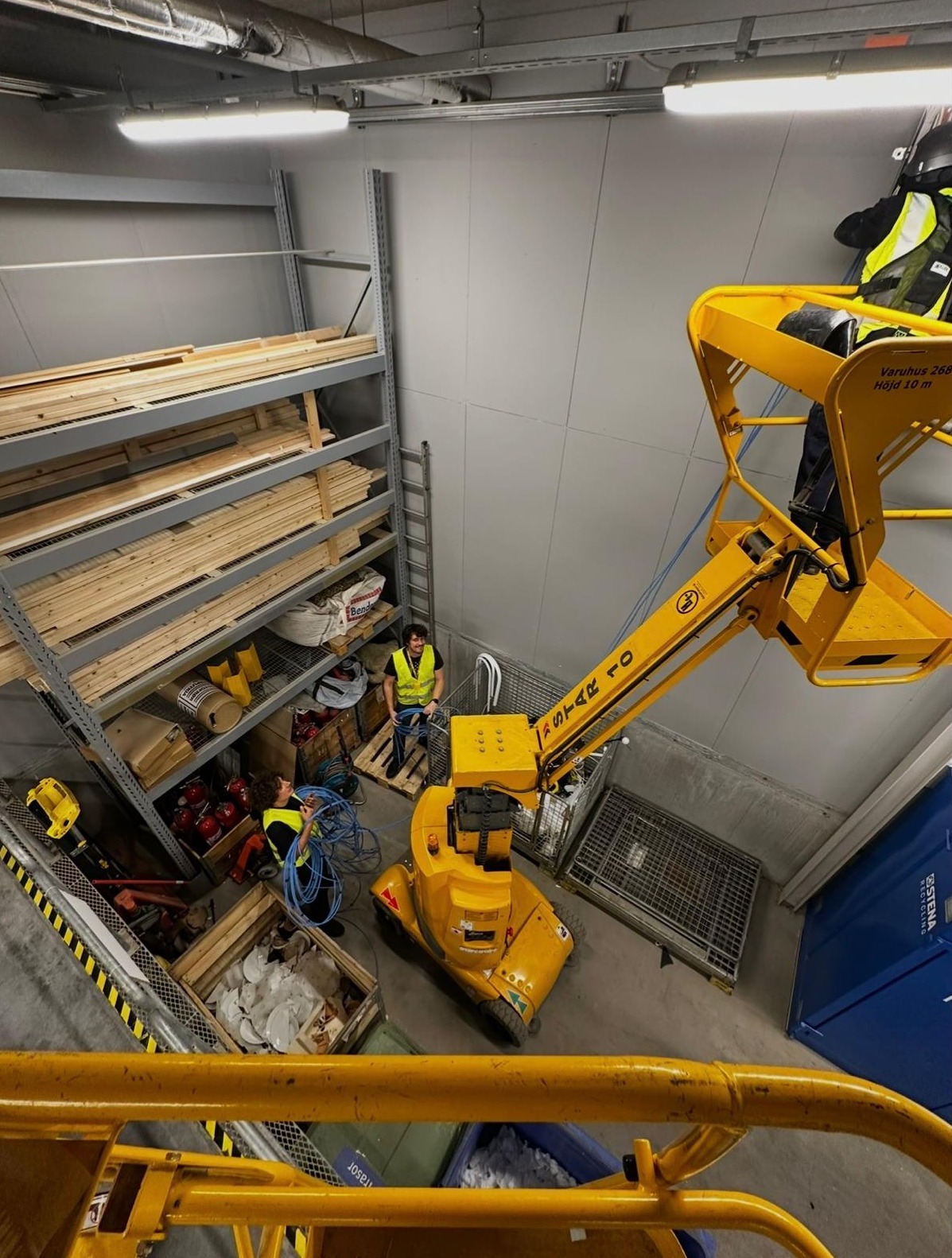 Praca na wysokości w magazynie: żółty podnośnik koszowy z pracownikami w kamizelkach odblaskowych, instalacja kabli przy regałach z drewnem. Widok z góry.