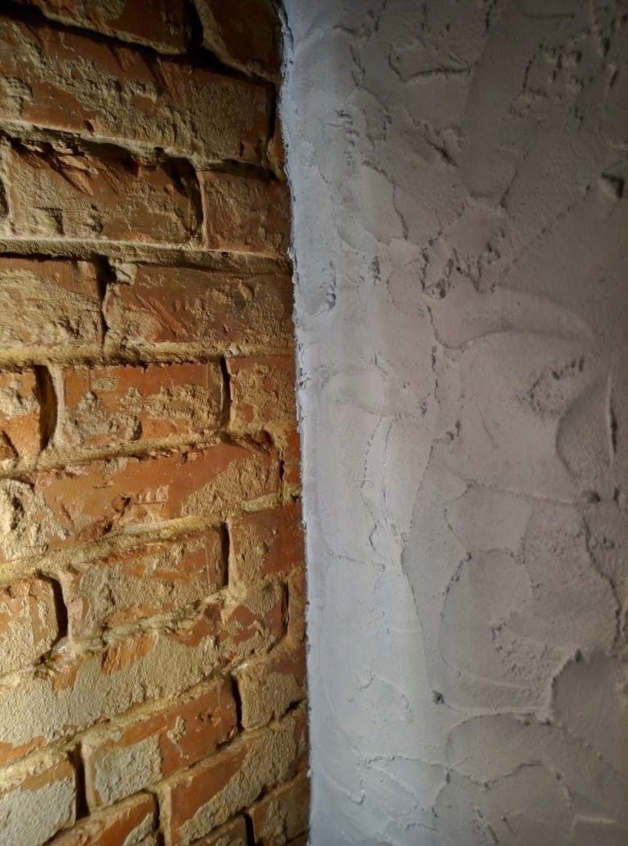 Fragment ściany: surowa cegła sąsiaduje z szarą, nierówną fakturą świeżo nałożonego tynku. Kontrast materiałów i faktur podkreślony przez boczne oświetlenie.