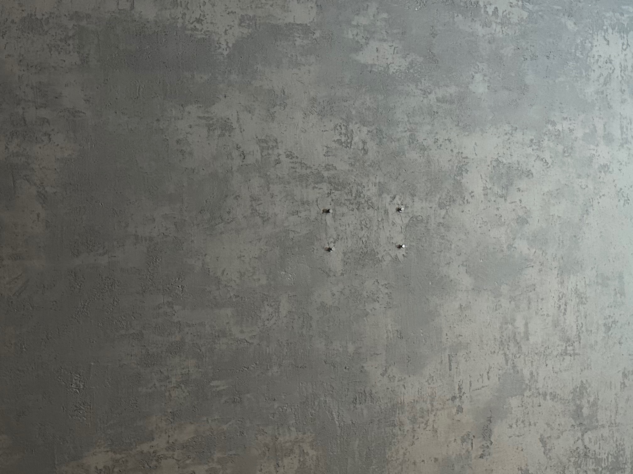 Szara ściana z efektem dekoracyjnym, imitującym beton. Widoczne cztery małe otwory po kołkach. Minimalistyczne wykończenie wnętrza.