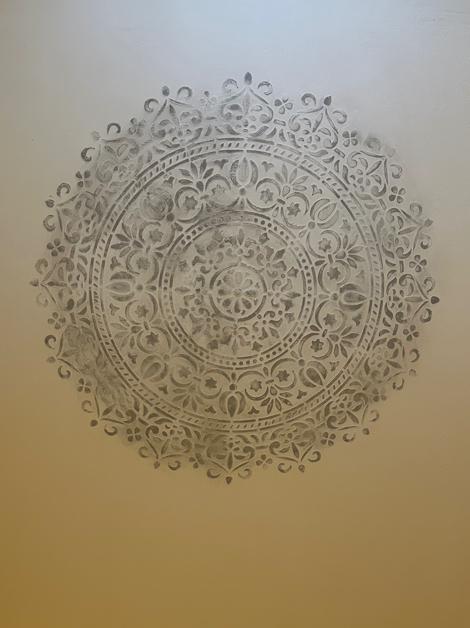 Dekoracyjne malowanie ściany z motywem mandali w odcieniach szarości. Wzór wykonany techniką szablonową, widoczny na gładkiej, jasnej powierzchni ściany.