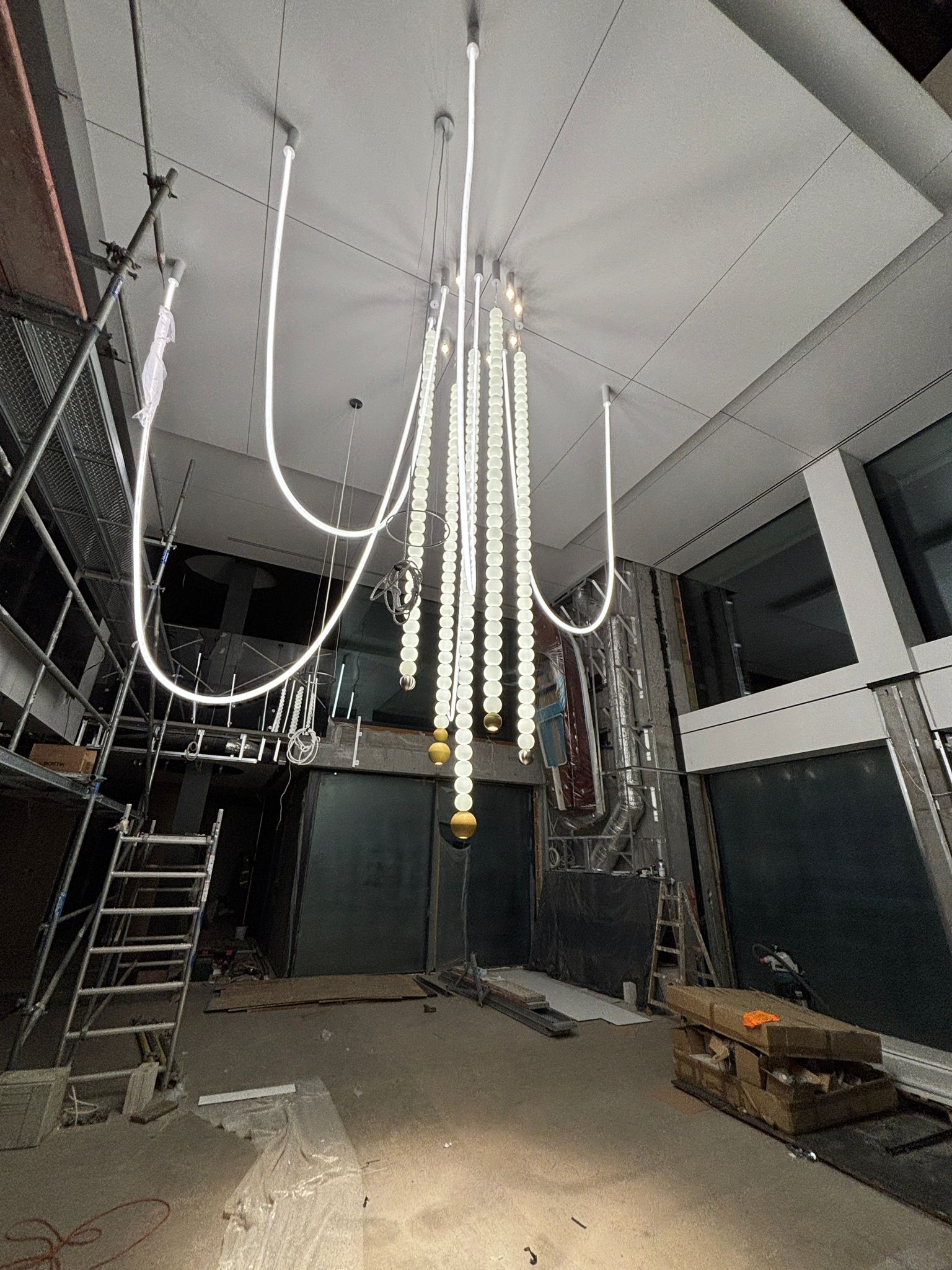 Nowoczesna lampa wisząca z elementami LED w surowym wnętrzu, widoczne rusztowania i elementy wykończeniowe. Nietypowy design oświetlenia w trakcie montażu.