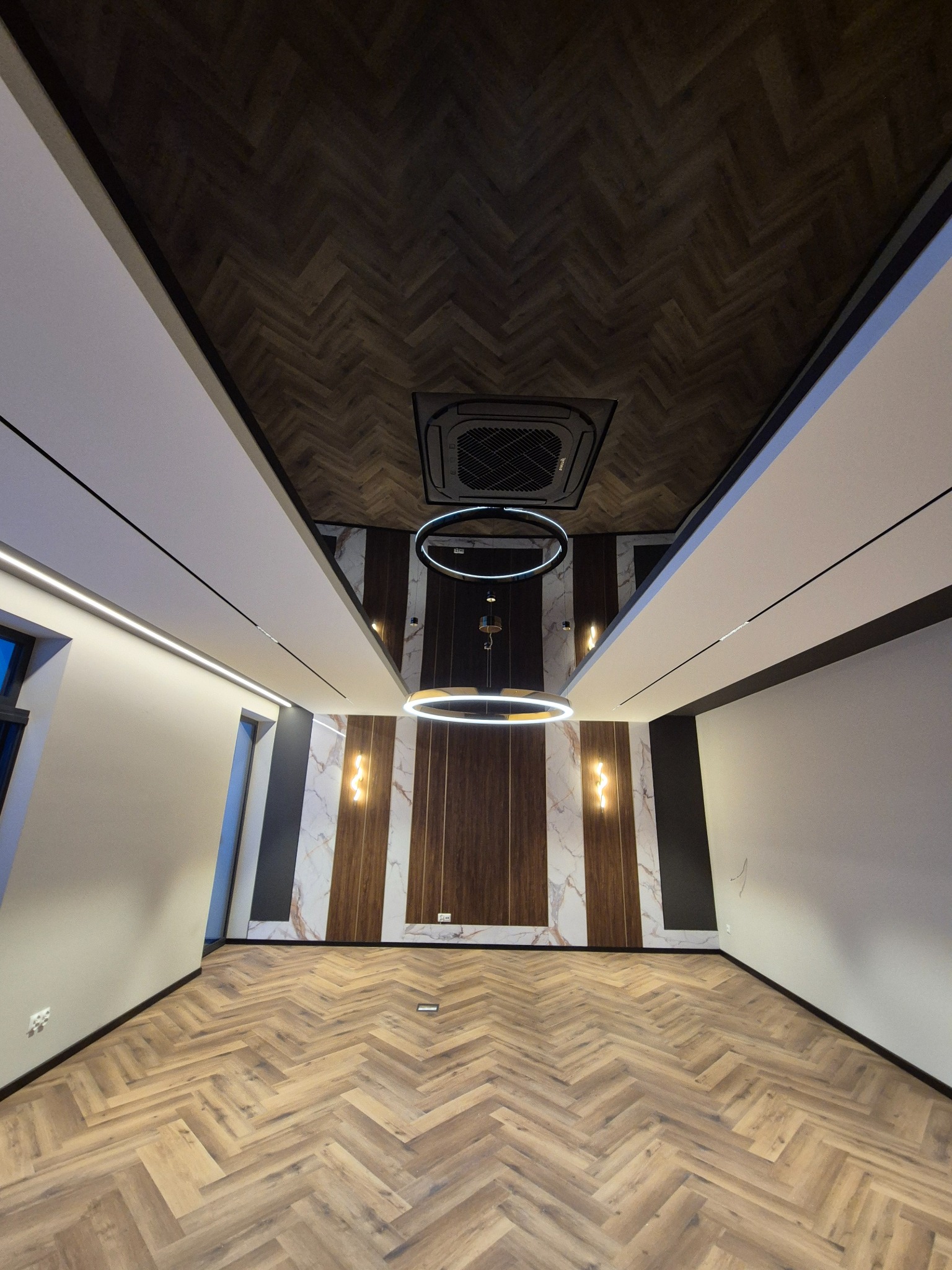 Nowoczesne wnętrze z parkietem w jodełkę, oświetleniem LED w suficie i okrągłą lampą wiszącą. Dekoracyjne panele ścienne z imitacją marmuru i drewna.