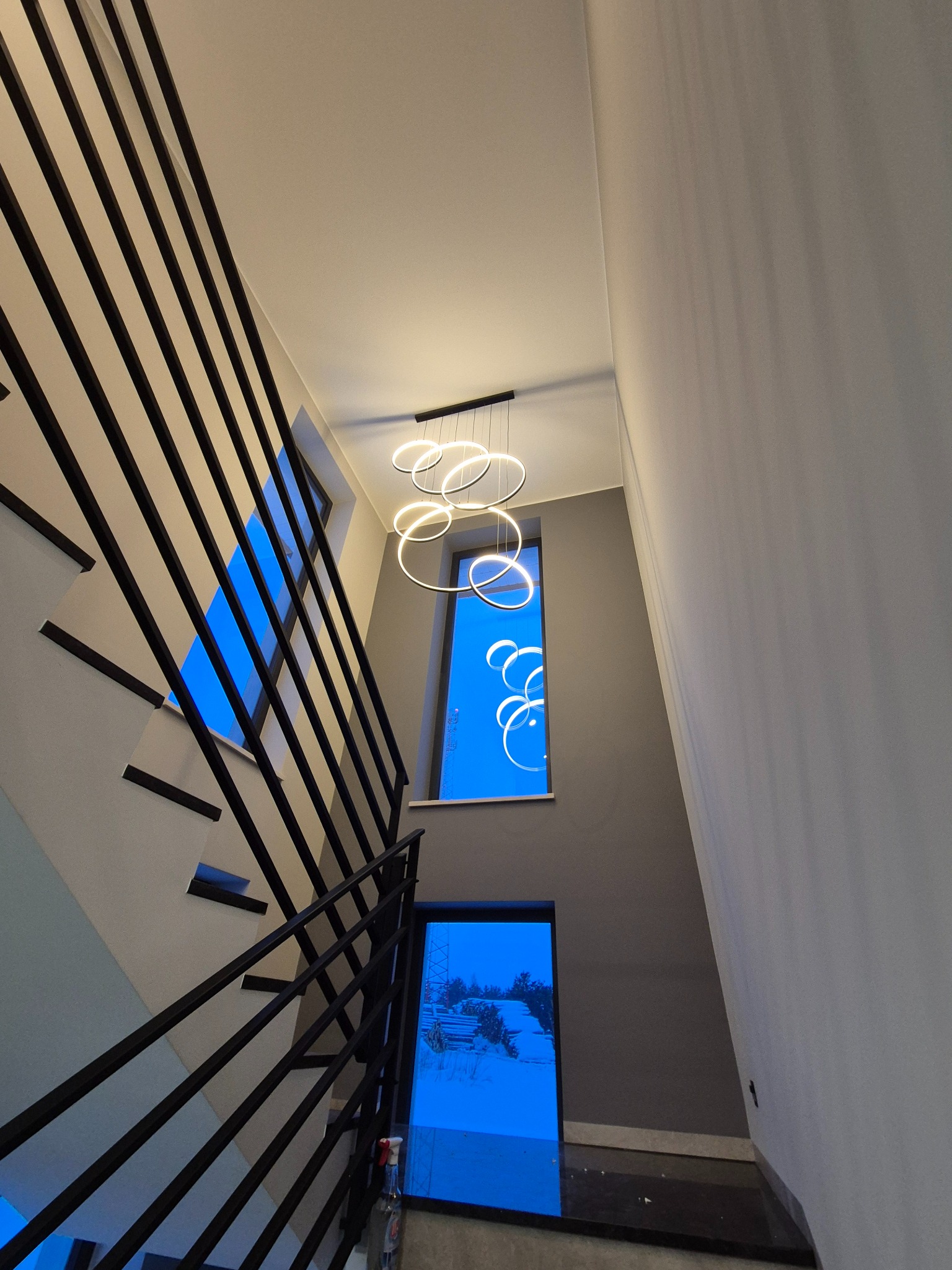 Nowoczesna lampa wisząca z okręgów LED oświetla klatkę schodową z czarną balustradą i oknami z widokiem na zewnątrz. Minimalistyczny design, jasne światło.