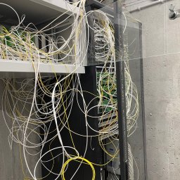 TEL-SOLID - Szafa rack z chaotycznie ułożonymi kablami światłowodowymi w kolorach białym i żółtym, widoczne elementy elektroniki i złącza. Betonowa ściana w tle.