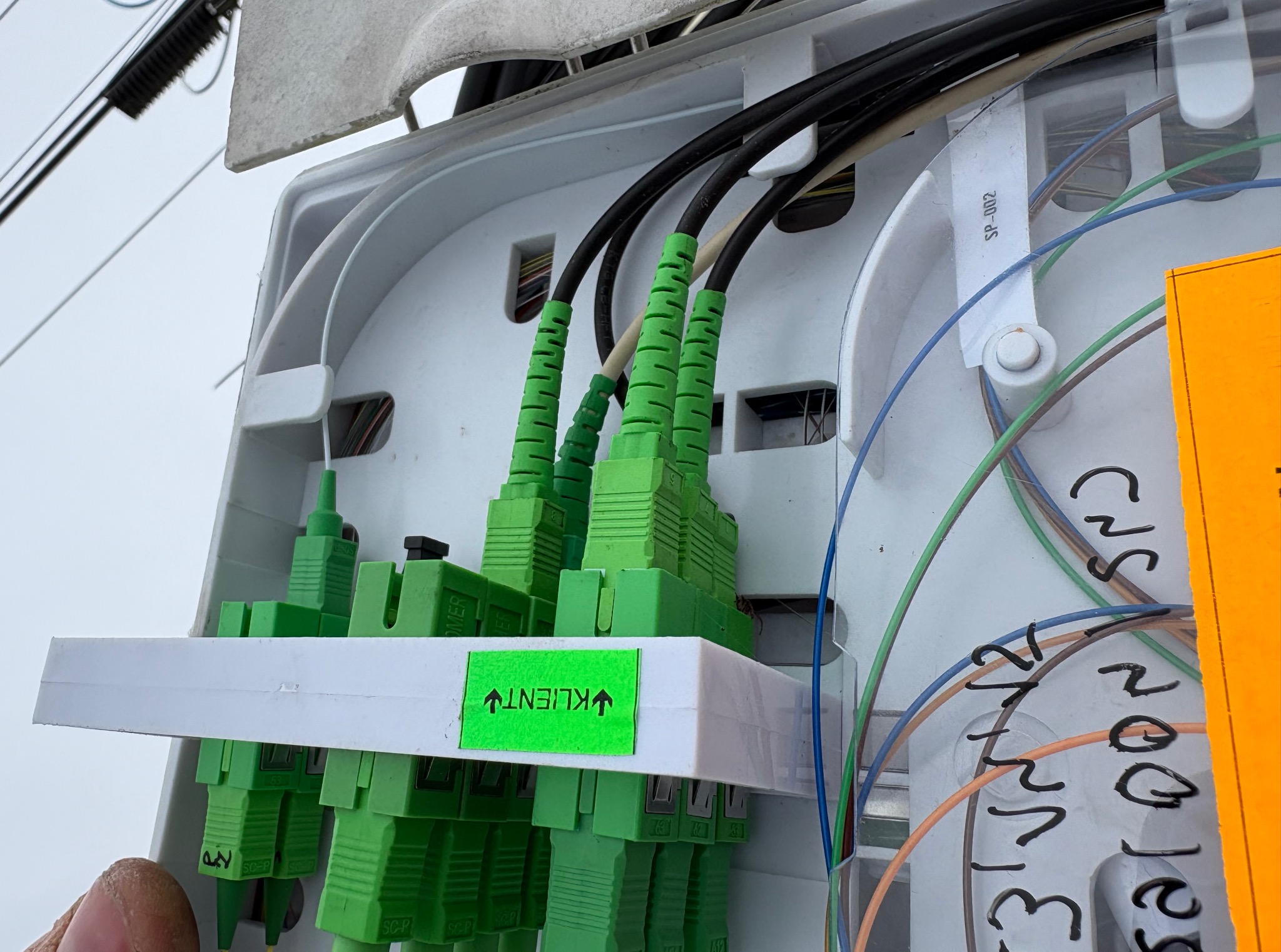Skrzynka telekomunikacyjna z podłączonymi światłowodami oznaczonymi zielonymi złączkami. Widoczna etykieta 'KLIENT' i oznaczenia kabli. Instalacja wewnątrz białej obudowy.