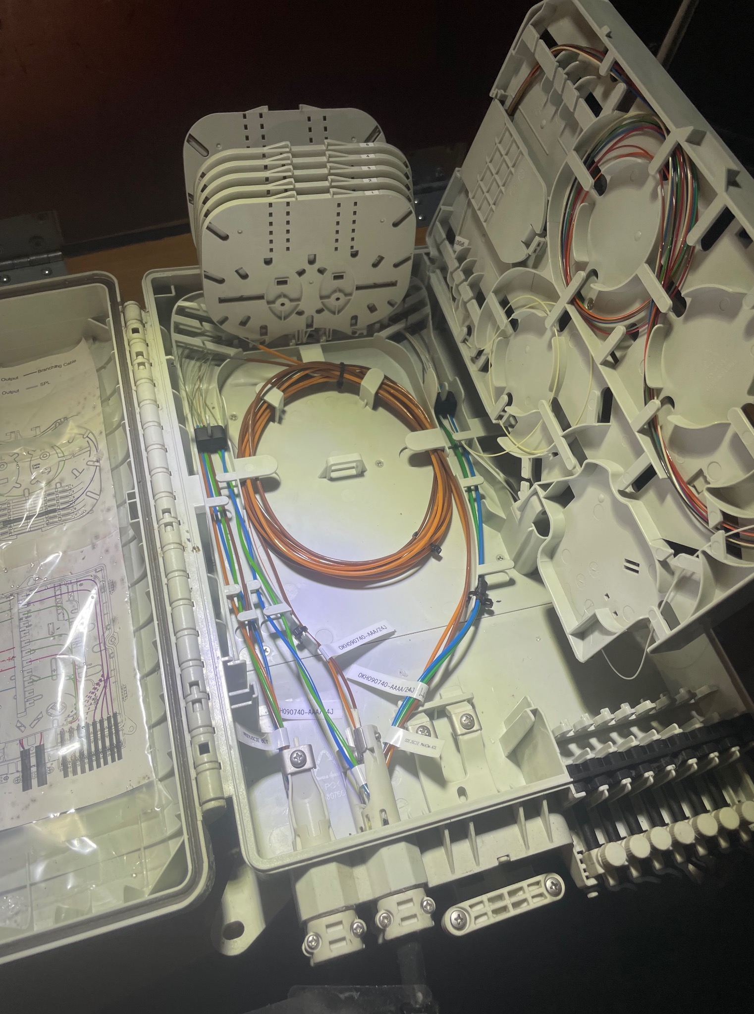 Otwarta skrzynka z kablami światłowodowymi wewnątrz, widoczne złącza, kable w różnych kolorach i schemat. Element instalacji telekomunikacyjnej w trakcie prac.