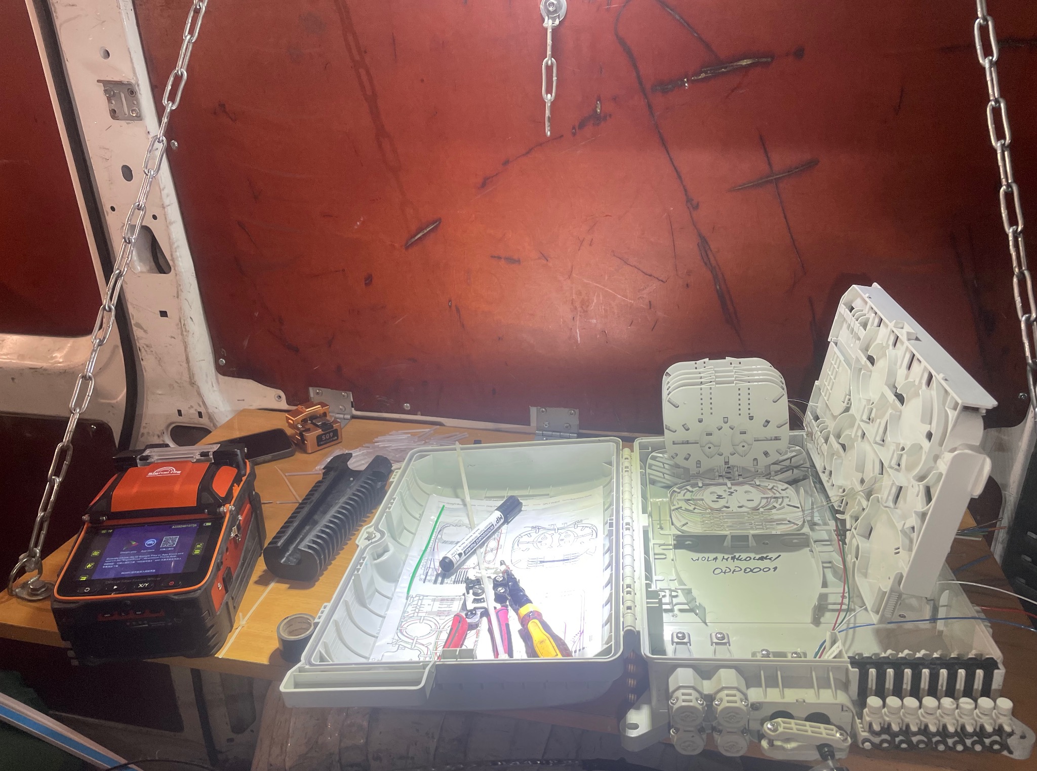 Otwarta skrzynka z narzędziami i schematem instalacji światłowodowej wewnątrz furgonetki, obok spawarka światłowodowa. Widoczne kable i złącza.