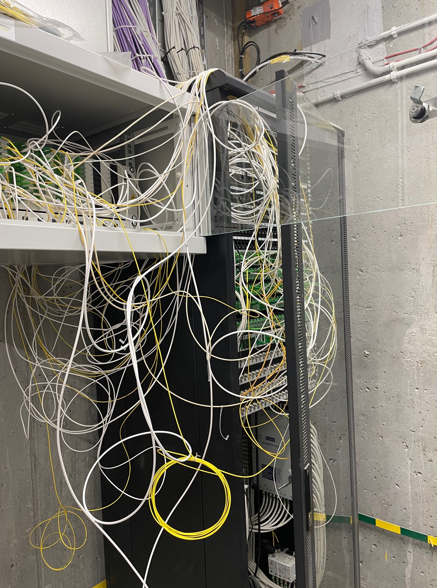 Szafa rack z chaotycznie ułożonymi kablami światłowodowymi w kolorach białym i żółtym, widoczne elementy elektroniki i złącza. Betonowa ściana w tle.