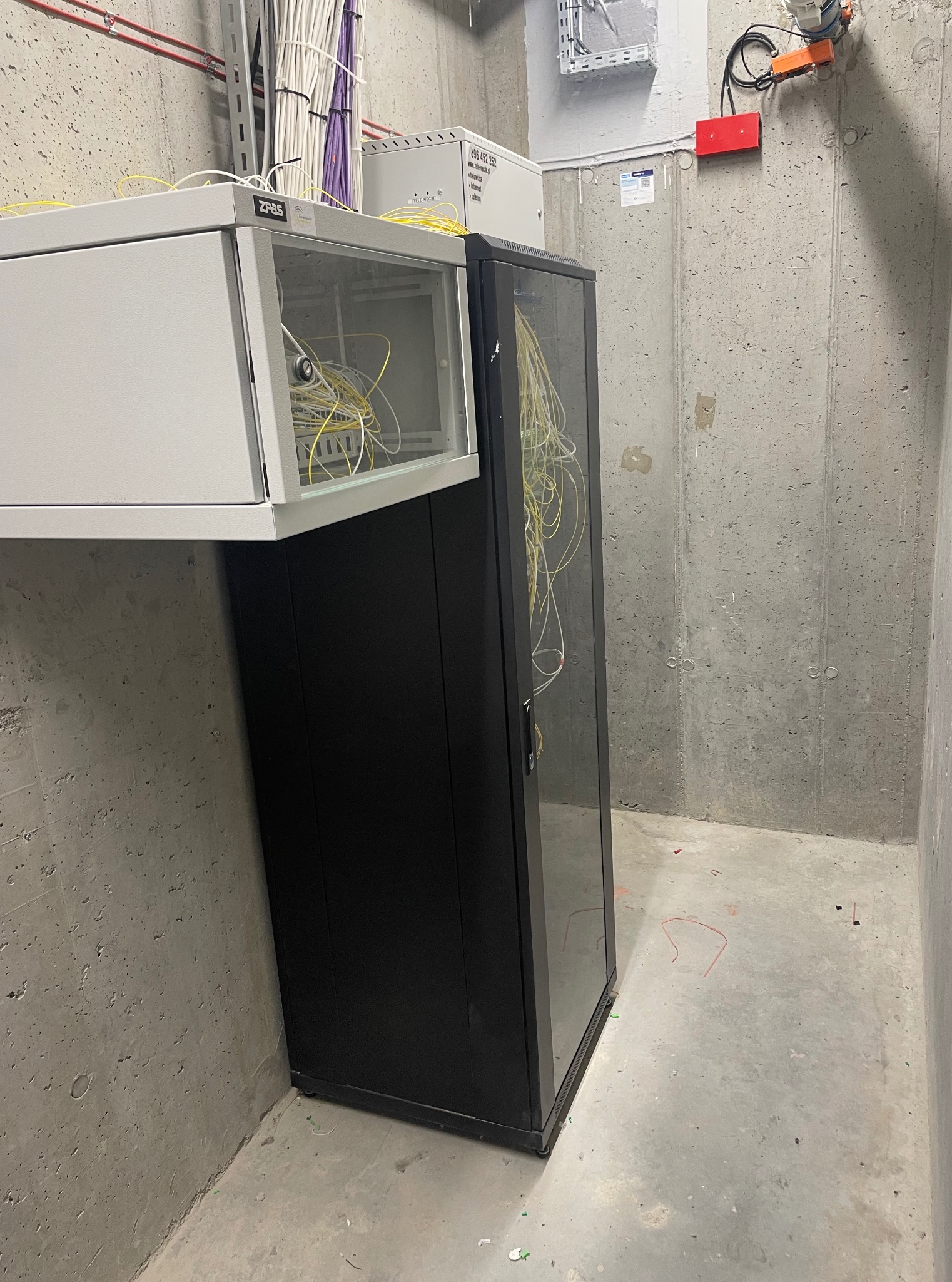 Szafa rack z okablowaniem w surowym pomieszczeniu. Widoczne kable w różnych kolorach. Betonowe ściany i podłoga. Górna szafka częściowo otwarta, dolna zamknięta.