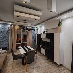 Nowoczesne wnętrze: szara ściana z kamienia dekoracyjnego, stół z krzesłami, czarno-biała szafka RTV i designerski sufit z oświetleniem punktowym. Całość w stonowanej kolorystyce.