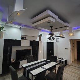 Nowoczesny salon z designerskim sufitem podwieszanym, oświetleniem LED i wentylatorem sufitowym. Minimalistyczne meble i dekoracyjna ściana z kamienia.