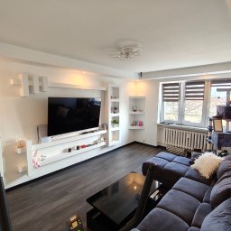 Nowoczesny salon z szarą sofą, czarnym stolikiem i ścianą TV z białymi półkami. Parkiet w odcieniach szarości. Widoczne okno z żaluzjami i drapak dla kota.