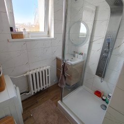 Jasna łazienka po remoncie: kabina prysznicowa ze srebrnym panelem, umywalka z szafką, białe płytki imitujące marmur, okno z widokiem na błękitne niebo.