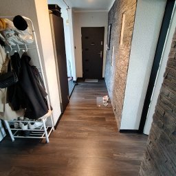 Konrad Warzyński - Wykończony korytarz z panelami podłogowymi imitującymi drewno i dekoracyjną ścianą z kamienia. Widoczny wieszak na ubrania i drzwi wejściowe.