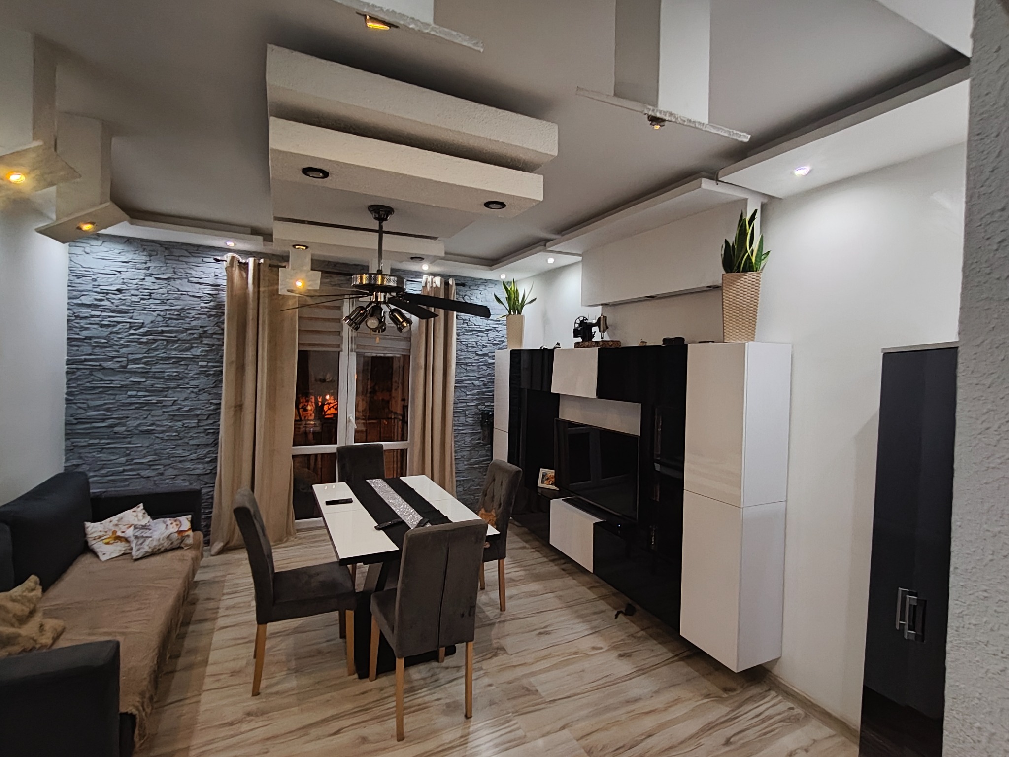 Nowoczesne wnętrze: szara ściana z kamienia dekoracyjnego, stół z krzesłami, czarno-biała szafka RTV i designerski sufit z oświetleniem punktowym. Całość w stonowanej kolorystyce.