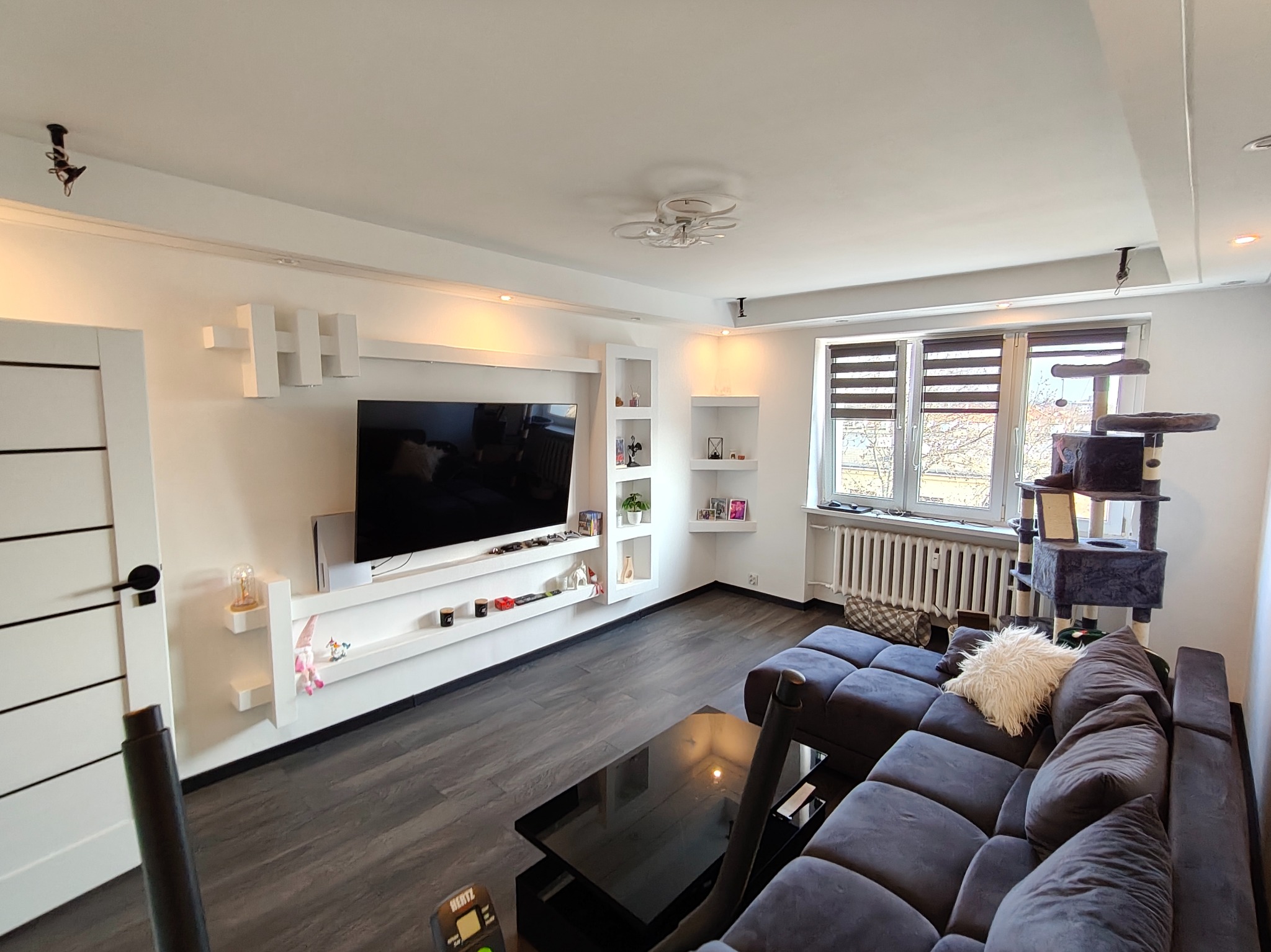 Nowoczesny salon z szarą sofą, czarnym stolikiem i ścianą TV z białymi półkami. Parkiet w odcieniach szarości. Widoczne okno z żaluzjami i drapak dla kota.