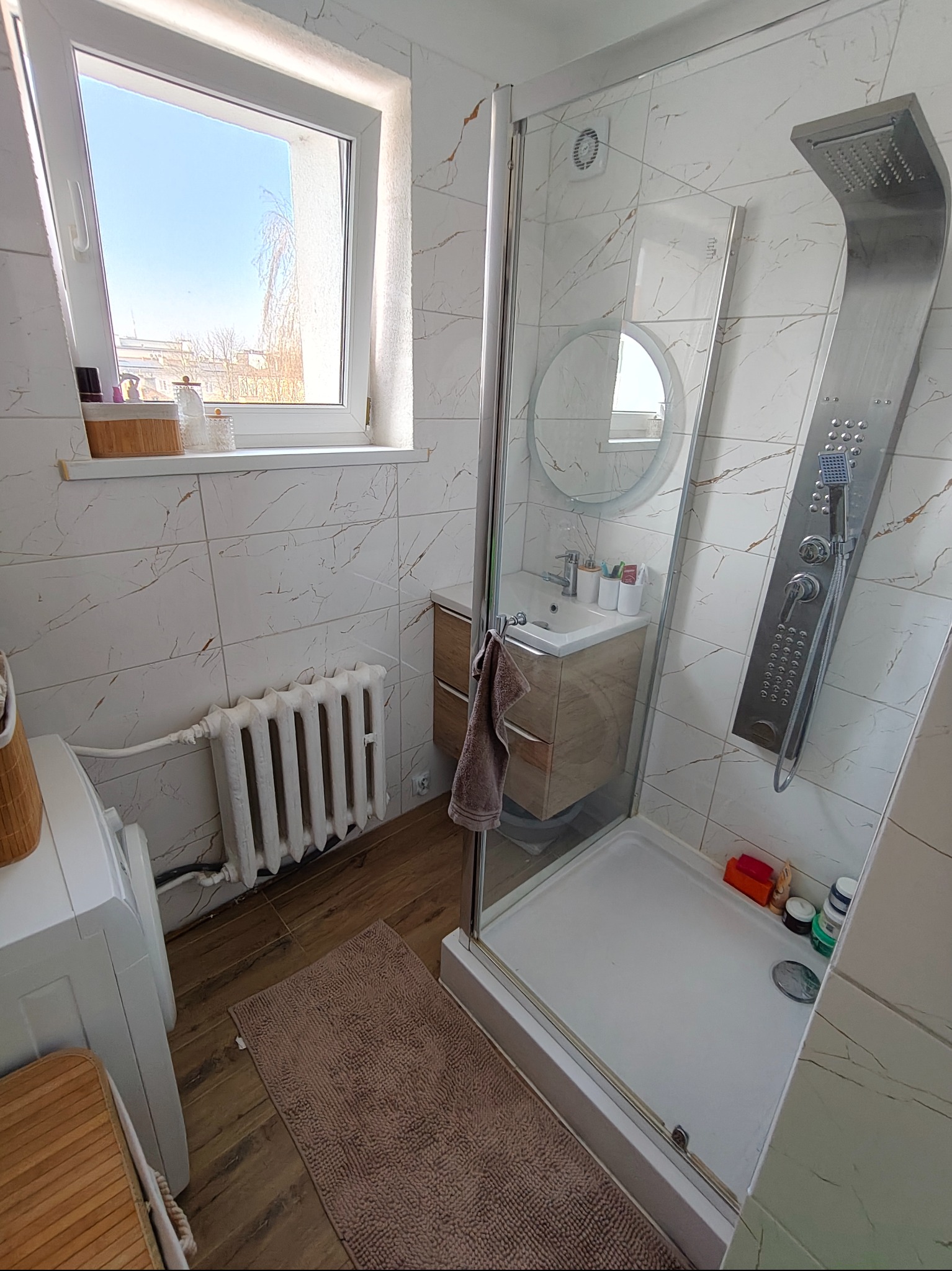 Jasna łazienka po remoncie: kabina prysznicowa ze srebrnym panelem, umywalka z szafką, białe płytki imitujące marmur, okno z widokiem na błękitne niebo.