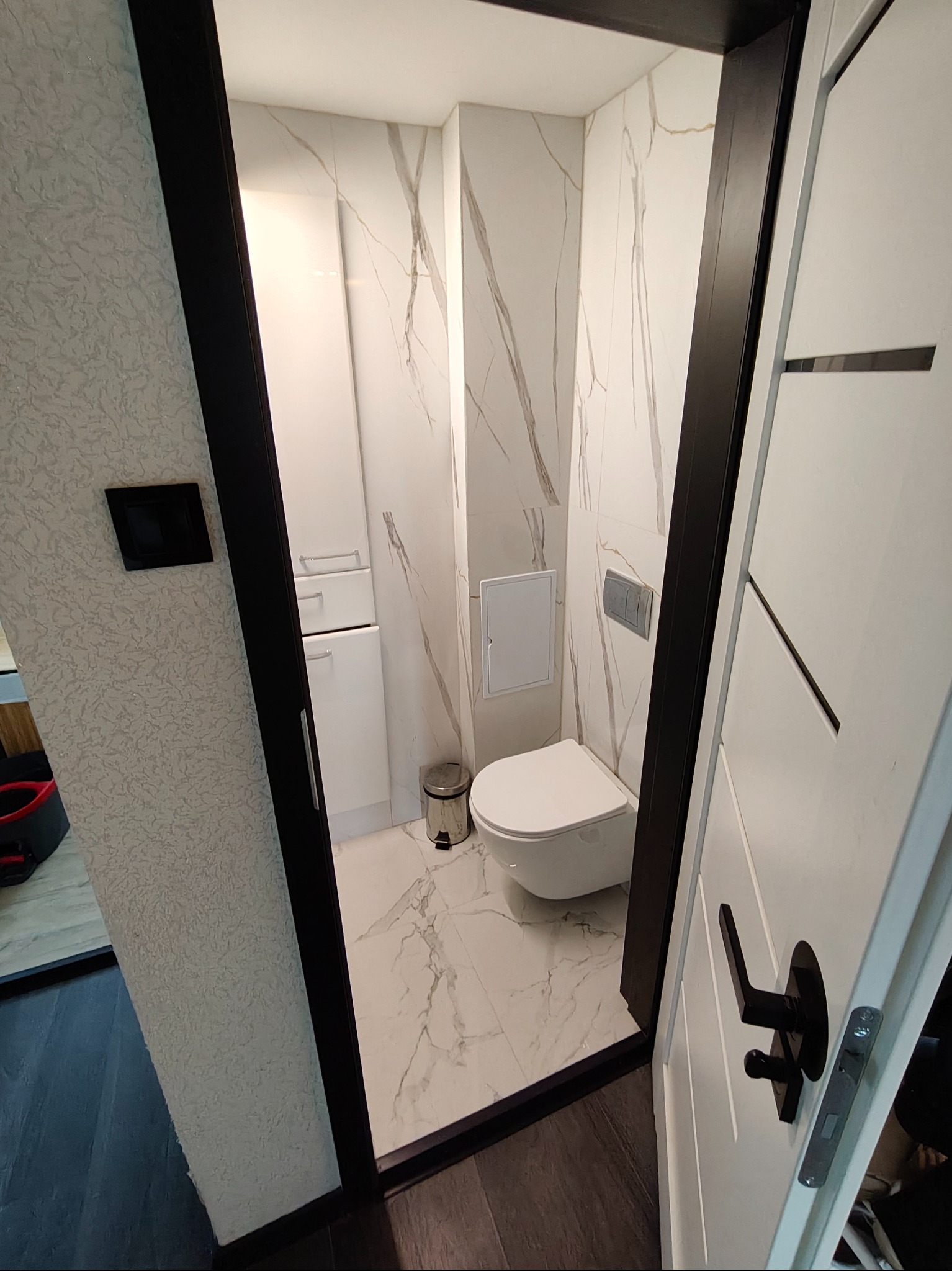 Wykończona łazienka z marmurowymi płytkami, białą armaturą i szafką w zabudowie. Widok zza uchylonych drzwi z czarną klamką. Nowoczesny design.