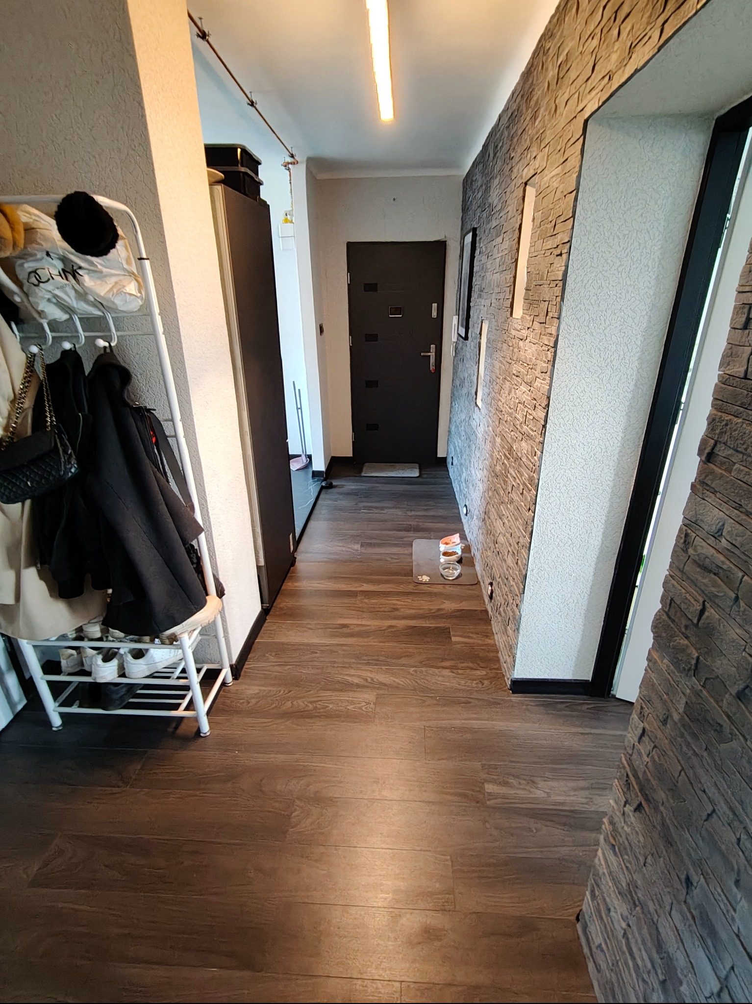 Wykończony korytarz z panelami podłogowymi imitującymi drewno i dekoracyjną ścianą z kamienia. Widoczny wieszak na ubrania i drzwi wejściowe.