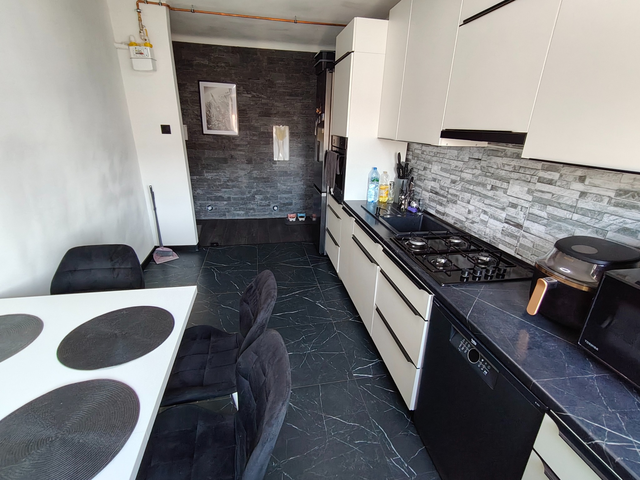 Nowoczesna kuchnia z białymi szafkami, czarnym blatem i marmurową podłogą. Ściana wykończona dekoracyjną cegłą. Widoczny stół z krzesłami.