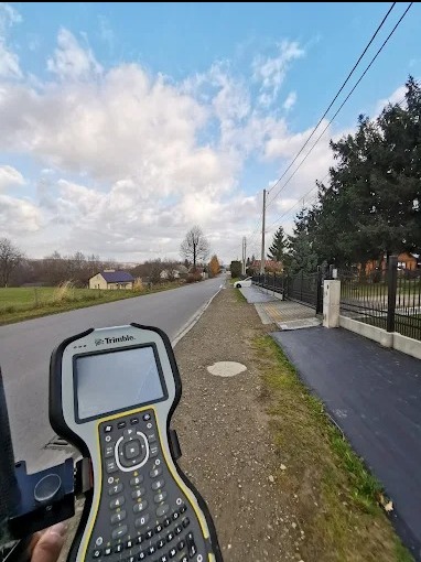 Geodeta w terenie z odbiornikiem GPS Trimble, mierzący działkę przy drodze i zabudowaniach. Widoczne niebo z chmurami.