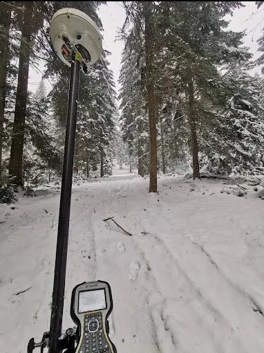 Praca geodezyjna w zimowym lesie: GPS na tyczce, ślady na śniegu, drzewa w tle. Pomiar terenu w trudnych warunkach pogodowych.