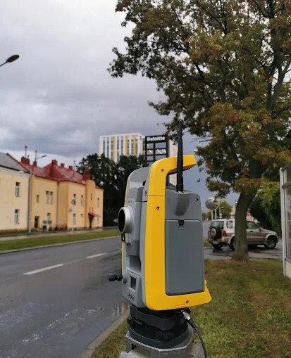 Żółto-szary teodolit na statywie, ustawiony na ulicy z budynkami w tle i pochmurnym niebem. Widoczny budynek Deloitte w oddali. Sprzęt geodezyjny w trakcie pracy.