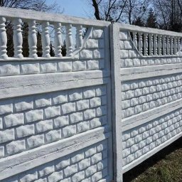 FACH KRZYŚ - Długi, biały, betonowy płot z dekoracyjnymi elementami, imitujący cegłę i balustrady, oddzielający teren. Widok z perspektywy, z zielenią w tle.