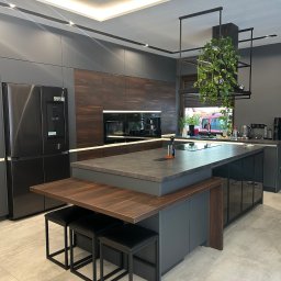 JB Service - Nowoczesna kuchnia z grafitowymi szafkami, drewnianymi panelami i blatem, dużą wyspą z płytą indukcyjną, smart lodówką i podświetleniem LED. Widoczna podwieszana roślina.