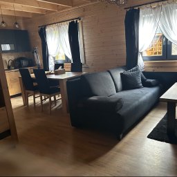 JB Service - Przytulne wnętrze drewnianego domu z otwartą przestrzenią: ciemna sofa, jasny stół jadalniany z krzesłami, nowoczesna kuchnia z ciemnymi szafkami. Widoczne drewniane ściany i belki sufitowe, okna...