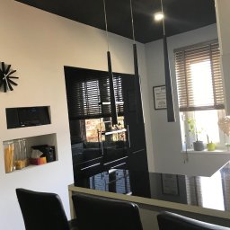 JB Service - Nowoczesna kuchnia z czarnym sufitem i białymi ścianami. Trzy podłużne lampy wiszą nad błyszczącym, czarnym blatem barowym z krzesłami. W tle okno z drewnianymi żaluzjami.