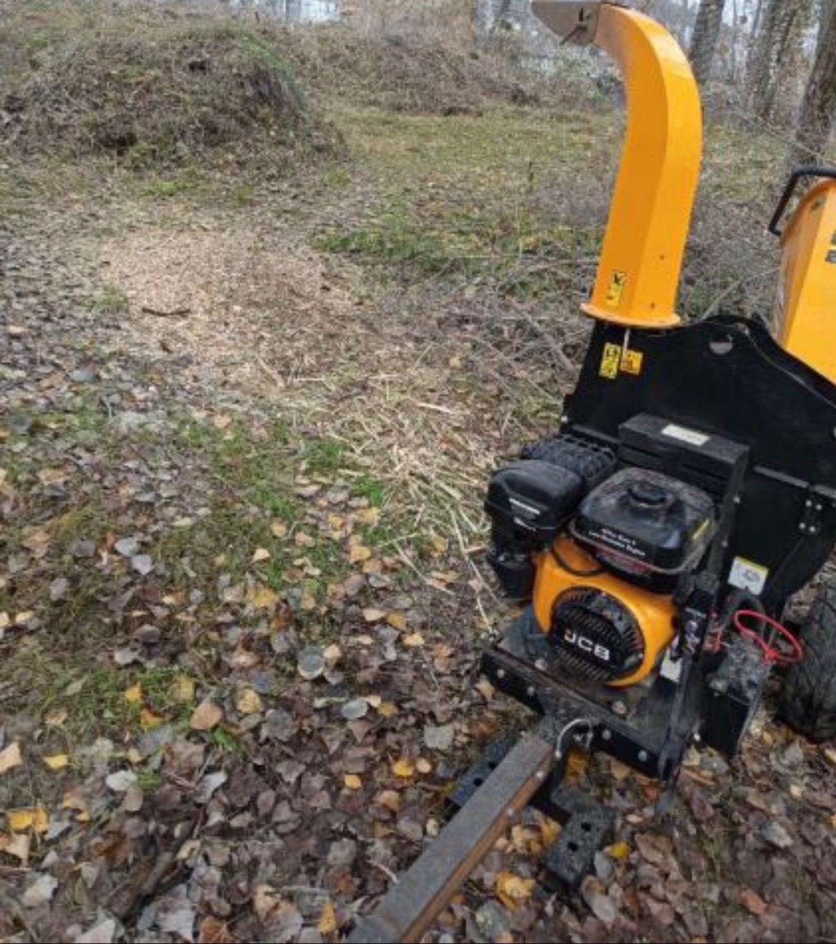 Żółto-czarny rębak do gałęzi JCB na tle trawy i liści, z widocznymi wiórami drzewnymi, sugerujący świeżo zakończone prace związane z wycinką lub pielęgnacją drzew.