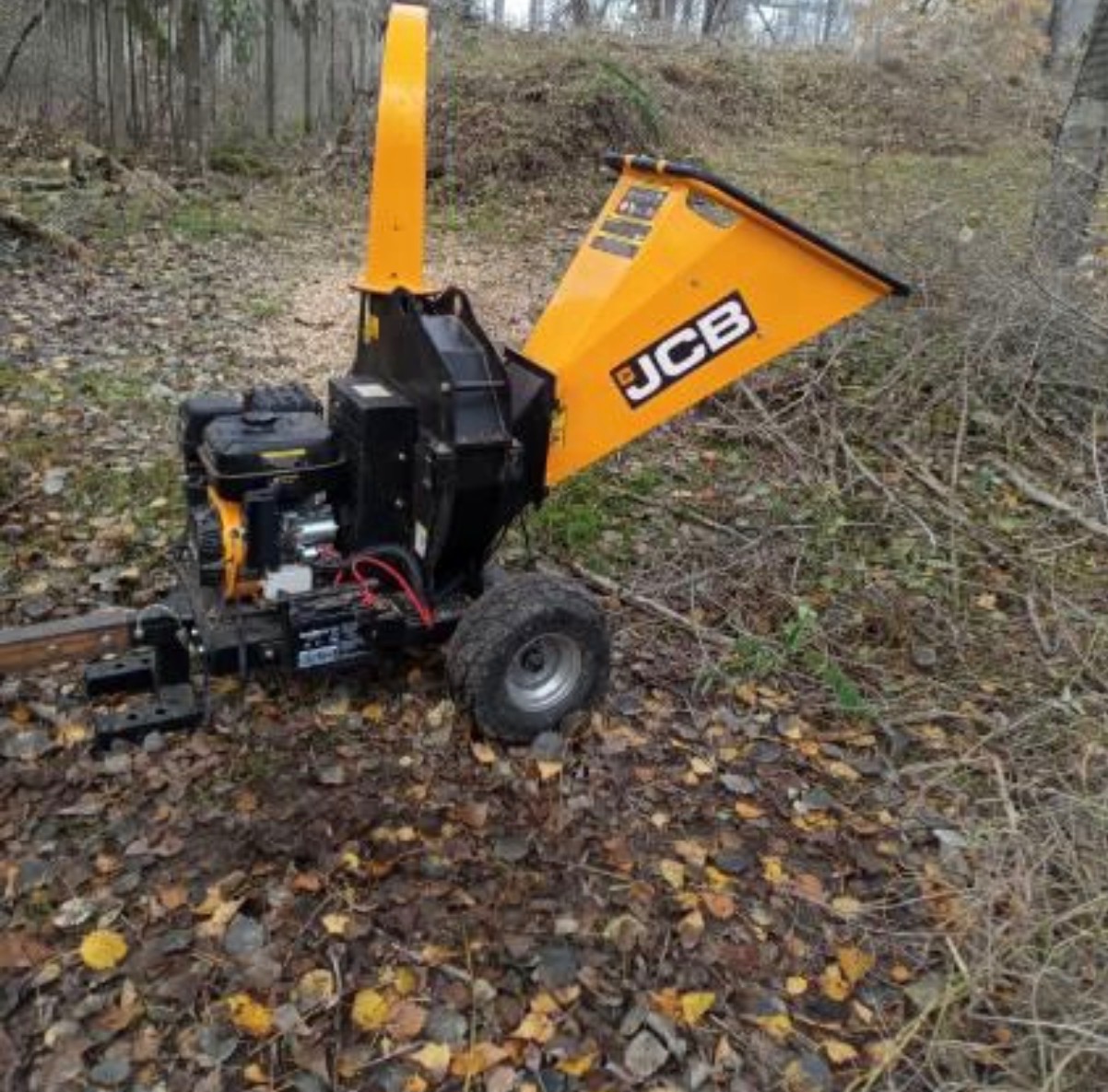 Żółto-czarny rębak do gałęzi JCB na tle opadłych liści i krzewów. Sprzęt gotowy do pracy, widoczne drobne gałęzie w pobliżu. Jesienna sceneria.