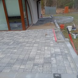 kacper łazarowicz - Układanie szarej kostki brukowej na tarasie domu z dużym oknem łukowym. Widoczne narzędzia, poziomica i worki z zaprawą. Częściowo ułożona nawierzchnia.