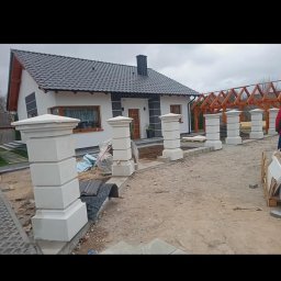 kacper łazarowicz - Nowoczesny dom z białymi ścianami i ciemną dachówką, otoczony dekoracyjnymi, kwadratowymi słupkami ogrodzeniowymi z jasnego kamienia.