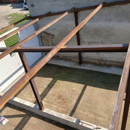 kacper łazarowicz - Konstrukcja stalowa dachu w trakcie budowy, widok z góry. Elementy zardzewiałej stali tworzą szkielet na tle ścian z pustaków i ziemi.