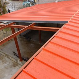 kacper łazarowicz - Czerwony dach z blachy trapezowej w trakcie montażu, widoczna stalowa konstrukcja nośna. Krople wody na powierzchni dachu. Widok z góry na budowę.