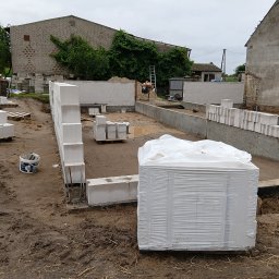 kacper łazarowicz - Budowa domu z betonu komórkowego: częściowo wymurowane ściany fundamentowe, paleta z bloczkami w folii, widoczny murarz przy pracy, budynek gospodarczy w tle.