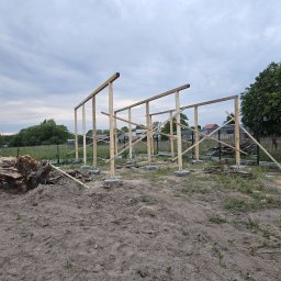kacper łazarowicz - Wznoszenie drewnianej konstrukcji szkieletowej na betonowych fundamentach, widoczne elementy z drewna i ziemia, w tle zieleń i zabudowania.
