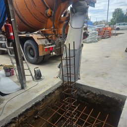 kacper łazarowicz - Betoniarka wylewa beton do wykopu zbrojonego stalą. Widoczna drabina, wiadro i zwinięty kabel. W tle budynki i błękitne niebo.