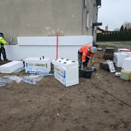 kacper łazarowicz - Ocieplanie fundamentów budynku styropianem IzoLbet. Dwóch pracowników w odblaskowych ubraniach przy pracy. Widoczne bloki styropianowe i narzędzia.