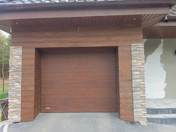 Brama garażowa segmentowa w odcieniach brązu, z kamienną okładziną po bokach, wkomponowana w elewację domu z drewnianą podbitką dachową i fragmentem otynkowanej ściany.