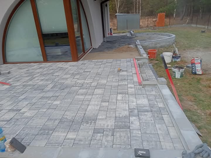 Układanie szarej kostki brukowej na tarasie domu z dużym oknem łukowym. Widoczne narzędzia, poziomica i worki z zaprawą. Częściowo ułożona nawierzchnia.