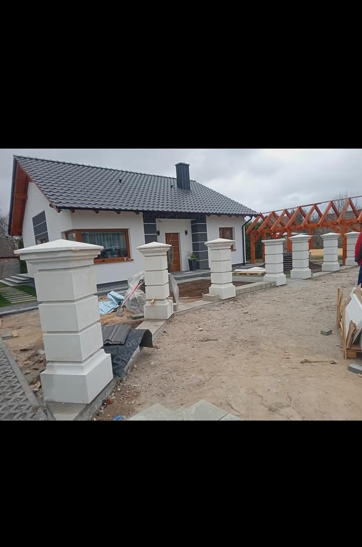 Nowoczesny dom z białymi ścianami i ciemną dachówką, otoczony dekoracyjnymi, kwadratowymi słupkami ogrodzeniowymi z jasnego kamienia.