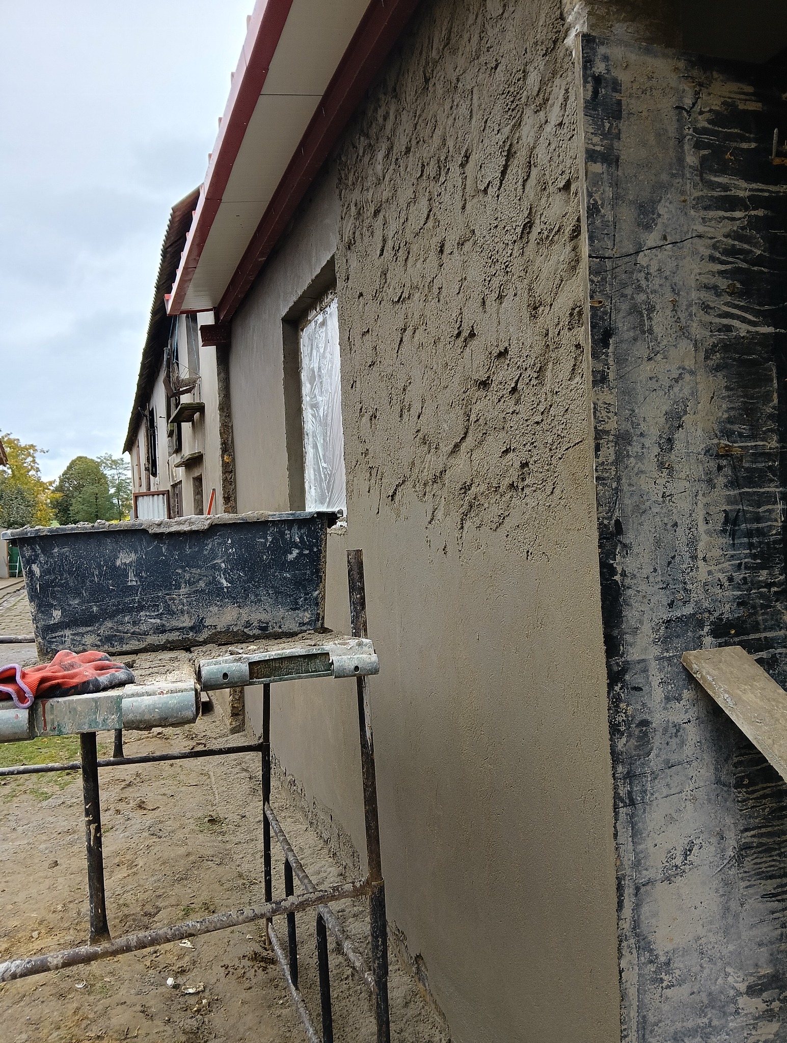 Ściana budynku w trakcie nakładania tynku zewnętrznego, widoczne rusztowanie z pojemnikiem i rękawicami roboczymi. Okno zabezpieczone folią.