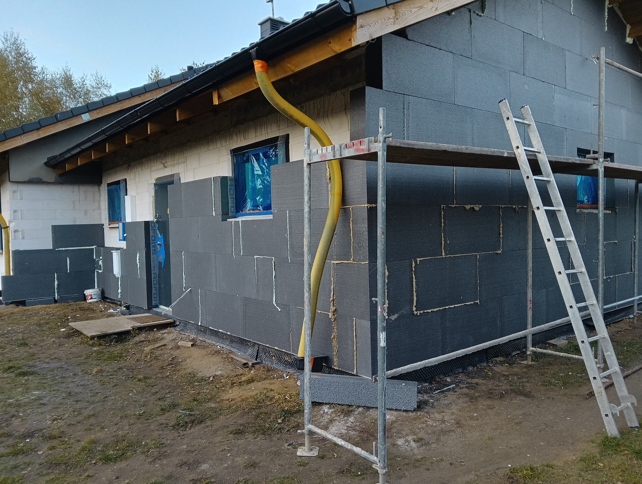Ocieplanie domu styropianem grafitowym, widoczne rusztowanie i żółty wąż wentylacyjny. Częściowo ocieplona ściana z białego bloczka. Okna zabezpieczone folią.