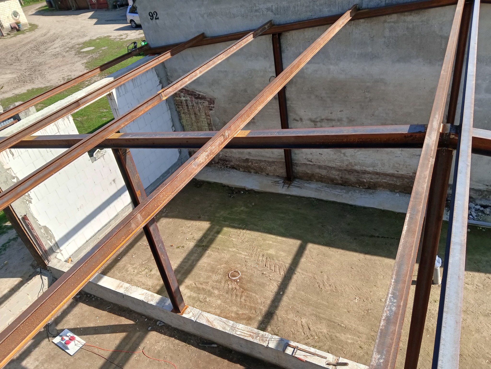 Konstrukcja stalowa dachu w trakcie budowy, widok z góry. Elementy zardzewiałej stali tworzą szkielet na tle ścian z pustaków i ziemi.