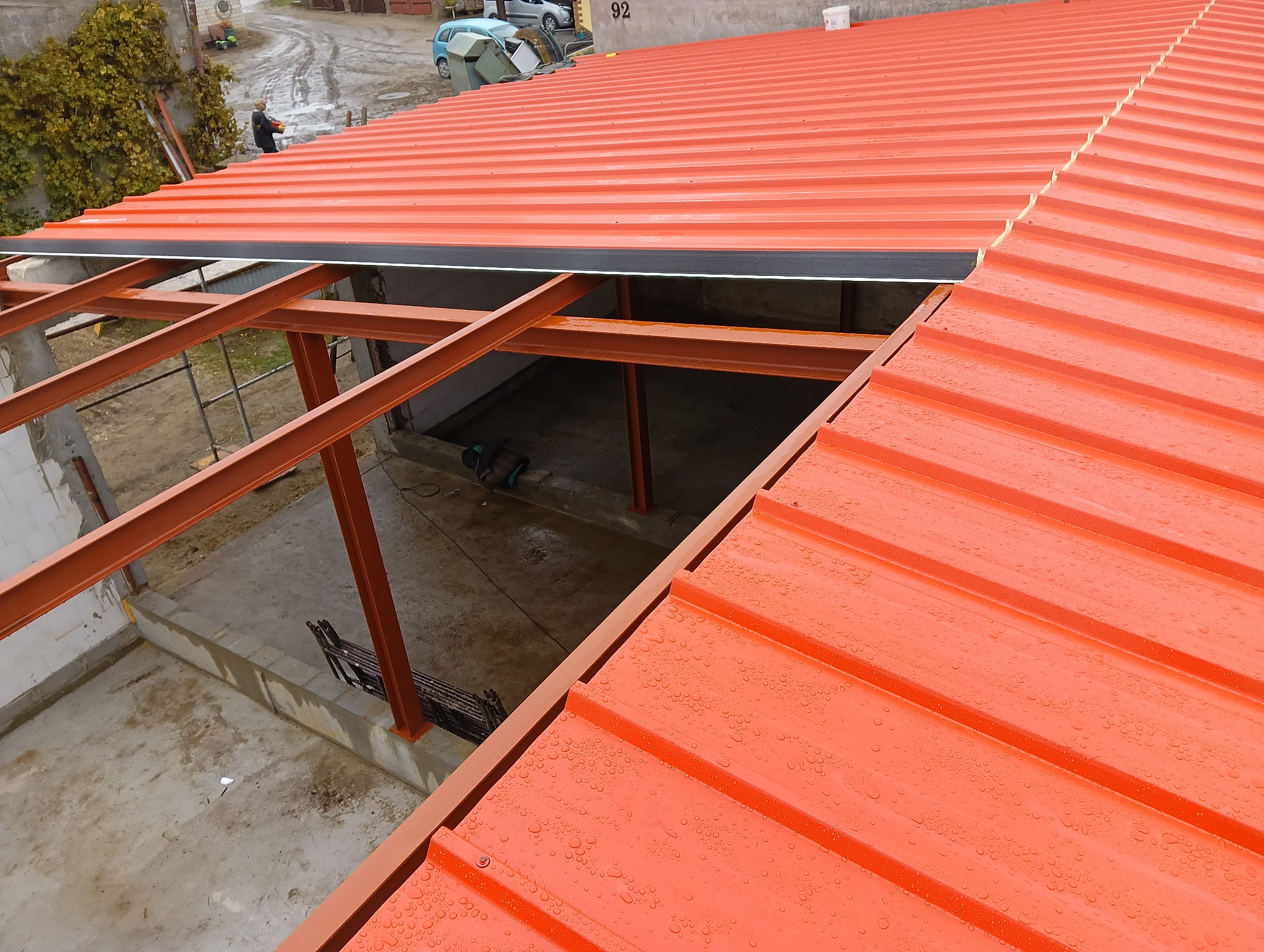 Czerwony dach z blachy trapezowej w trakcie montażu, widoczna stalowa konstrukcja nośna. Krople wody na powierzchni dachu. Widok z góry na budowę.